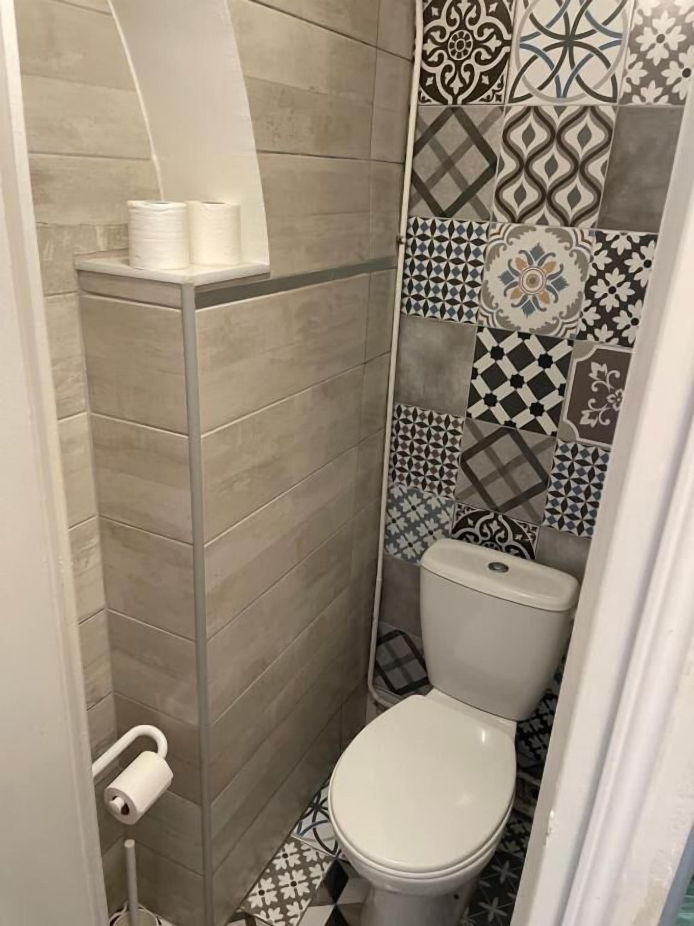 A small, tiled bathroom with a toilet, a roll of toilet paper, and a shower area.