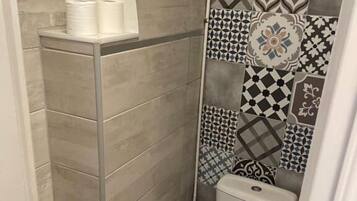 A small, tiled bathroom with a toilet, a roll of toilet paper, and a shower area.