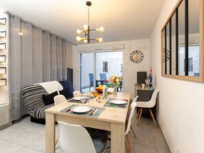 Un coin repas avec une table en bois, des chaises blanches et un lustre. On y trouve un canapé avec un coussin à rayures et une fenêtre avec des rideaux.