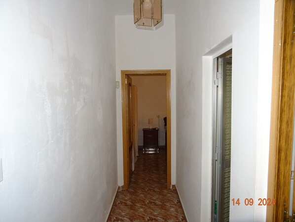 A narrow hallway with white walls and a wooden door on the right. A small table with a lamp is visible in the background.