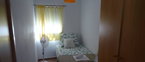 A small room with a bed, a standing fan, a window with curtains, and a wooden door.