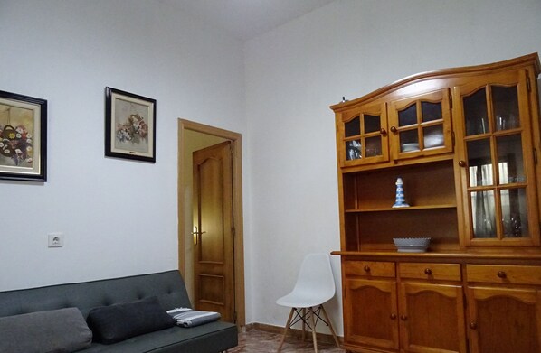 Un salón con un sofá verde oscuro, dos cuadros enmarcados con motivos florales, un mueble de madera con puertas de cristal y una silla blanca.