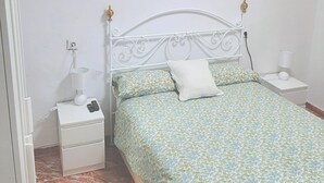 Een slaapkamer met een wit metalen bedframe, een bed met een patroon, een wit kussen, een bedlampje en een nachtkastje.