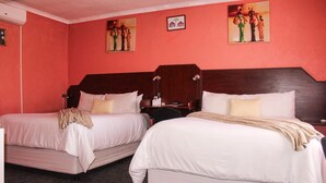 Camera d'albergo con due letti singoli, ognuno con lenzuola bianche e una coperta beige. Sono presenti due comodini con lampade e un tavolino tra i letti. Le pareti sono dipinte di rosso e ci sono due opere d'arte incorniciate sopra i letti.