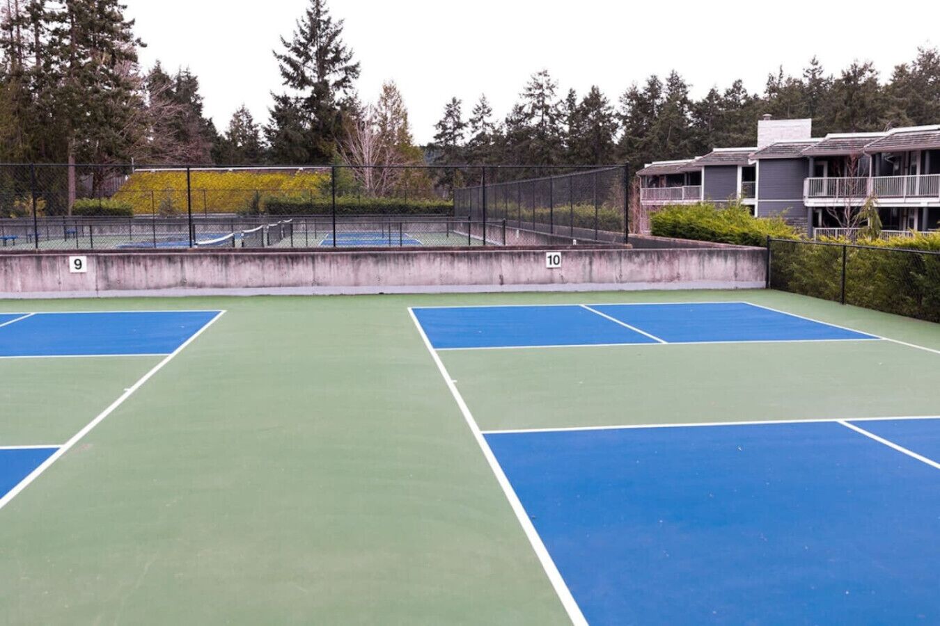 Un court de tennis extérieur avec des surfaces vertes et bleues, entouré d’une clôture et d’arbres.