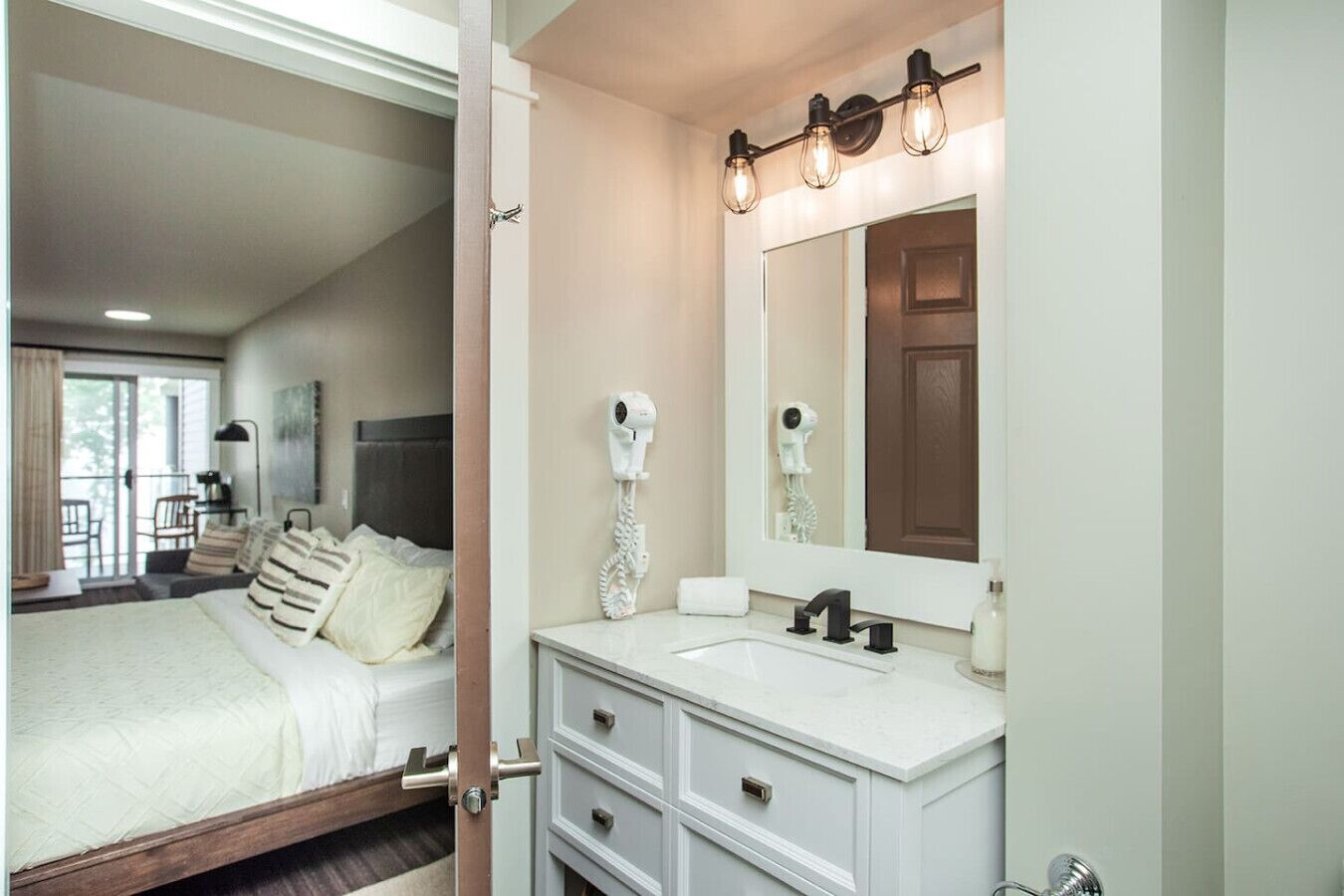 A hotel room with a bed, a white vanity with a mirror, and a hairdryer on the wall.