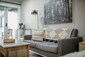 A modern living room with a grey sofa, a coffee table, and a bookshelf. - Forest Stream Escape Suite B (Nanoose Bay)
