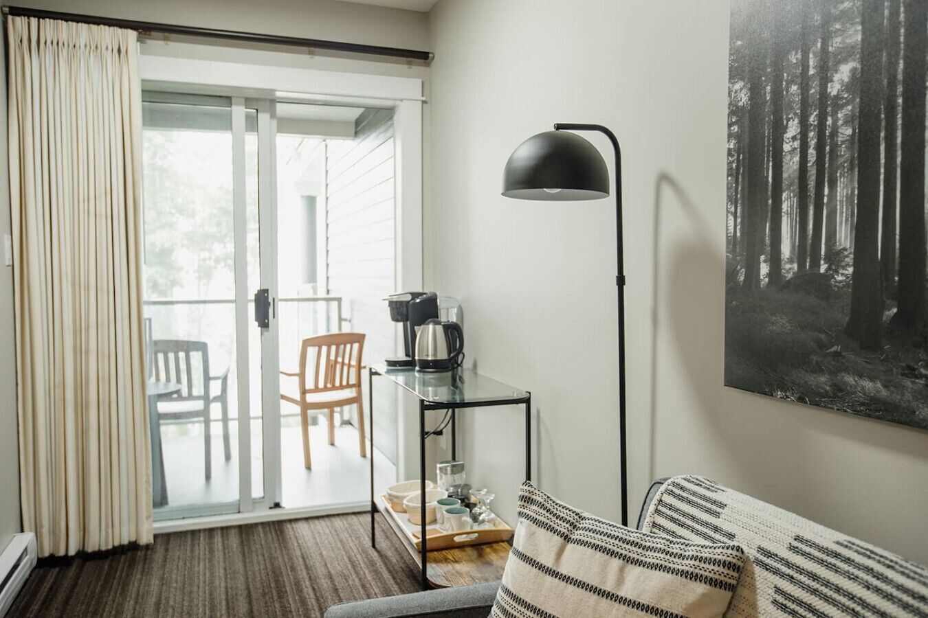 A modern living room with a glass door leading to a balcony, a coffee table with a kettle, a floor lamp, and a large forest-themed painting on the wall.
