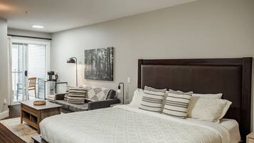 A bedroom with a large bed, a bedside table, a chair, a lamp, and a window with blinds.