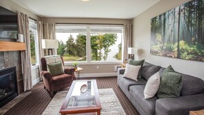 A living room with a fireplace, a sofa, a chair, a coffee table, and a large window with a forest view. - Escape & Unwind in Suite A (Nanoose Bay)