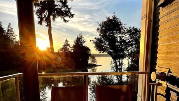 A wooden deck with two chairs and a table, overlooking a body of water and trees, with the sun setting in the background. - Escape & Unwind in Suite A (Nanoose Bay)
