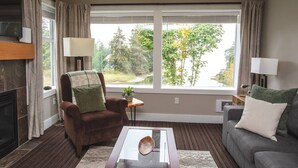 A living room with a fireplace, a brown armchair with a green pillow, a gray sofa with a white pillow, a coffee table, and a view of trees through large windows. - Escape & Unwind in Suite A (Nanoose Bay)