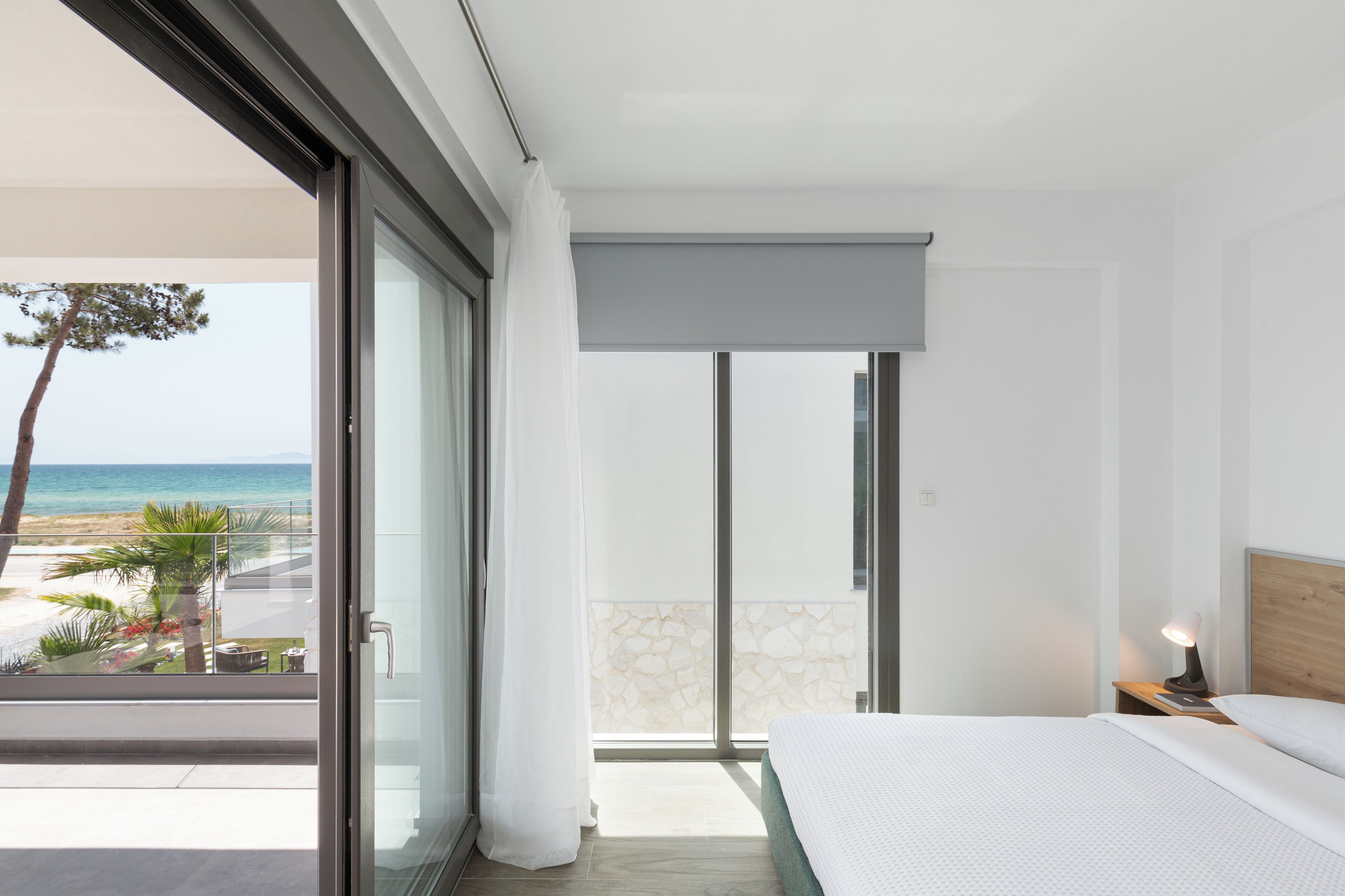 Une chambre d’hôtel moderne avec un balcon donnant sur une plage, un lit avec une tête de lit en bois et une table de chevet avec une lampe.