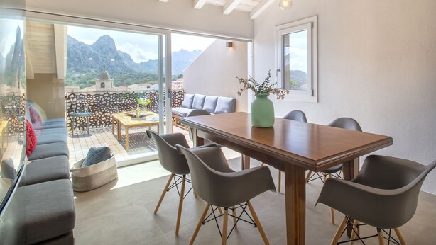 Un comedor moderno con una mesa y sillas de madera, un sofá y vistas a montañas a través de grandes ventanales.