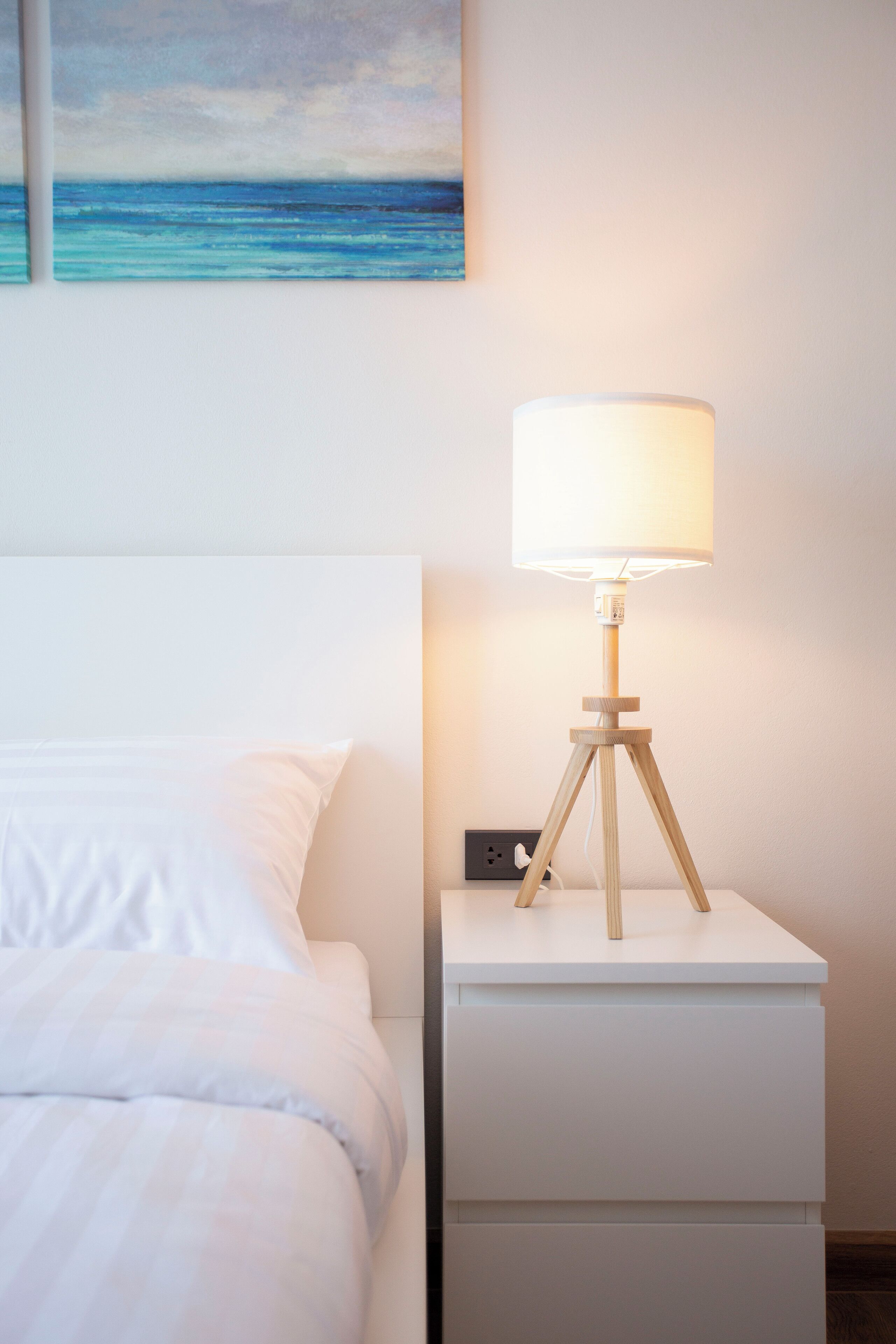 A neatly made bed with white linens, a bedside table with a lamp, and a painting of a blue ocean on the wall.