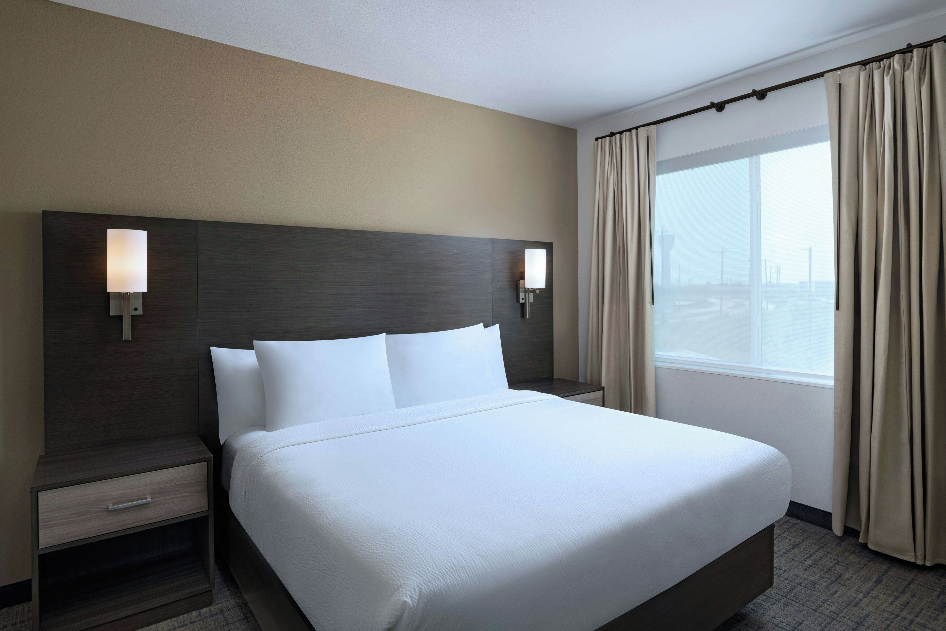 A hotel room with a large bed, two bedside tables, a headboard with wall sconces, a window with curtains, and a view of a cityscape.