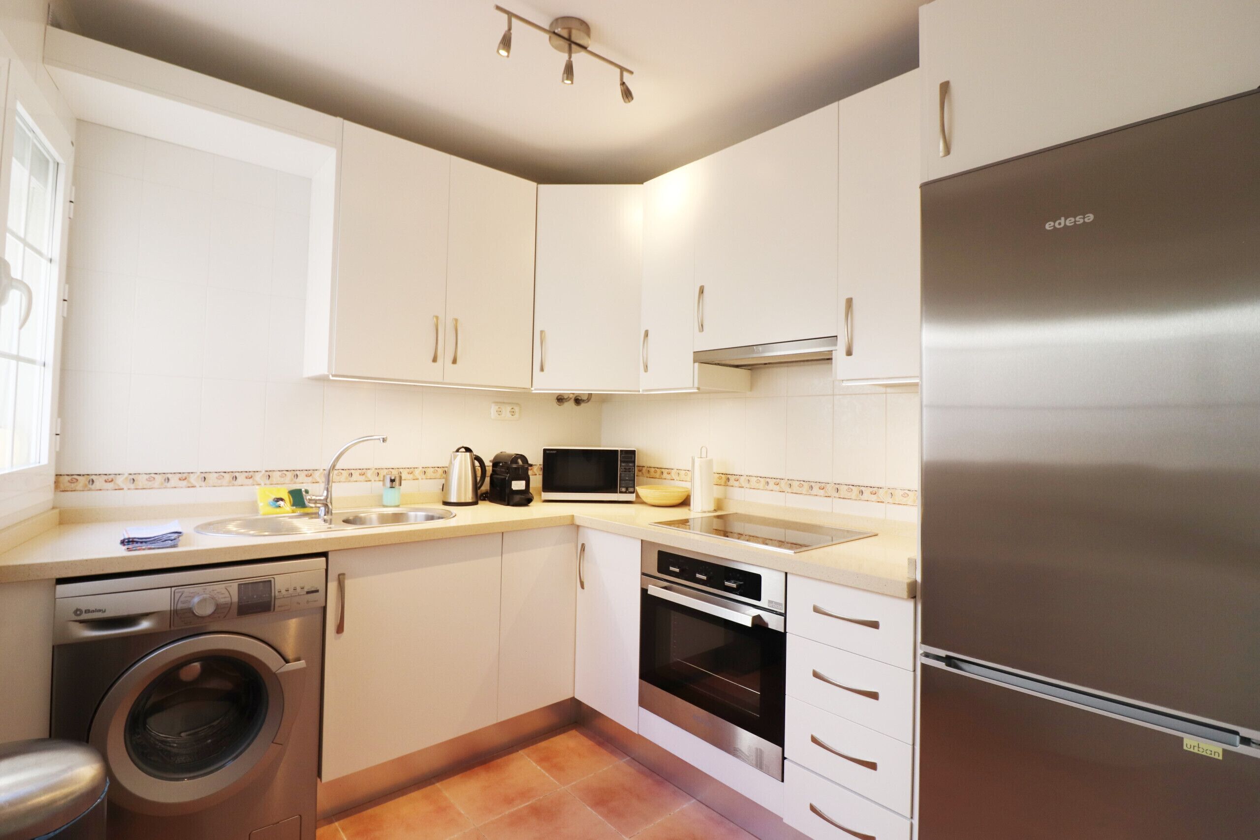 A compact kitchen with white cabinets, a stainless steel refrigerator, a washing machine, and a microwave.