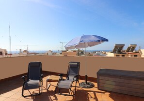 A rooftop terrace with two lounge chairs, a table, and a large umbrella providing shade.