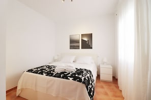 Un dormitorio con cama, cortinas blancas, una mesita de noche y dos cuadros colgados en la pared.