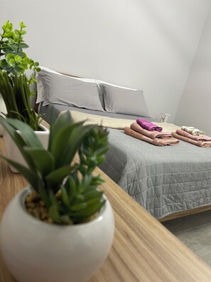 A neatly made bed with a grey quilt, white pillows, and folded towels in shades of pink and beige. There are potted plants on the bedside table.