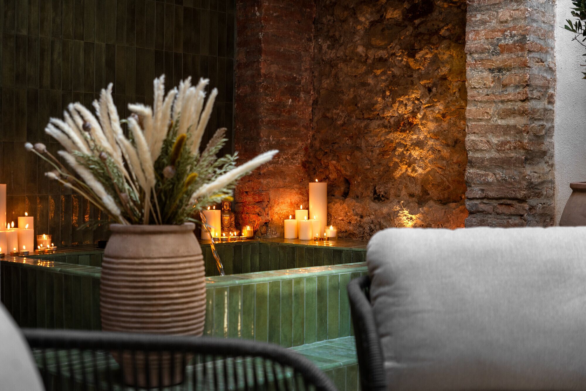 A cozy seating area with a green cushioned bench and a wicker chair, a vase with dried plants, and multiple lit candles on a brick wall backdrop.