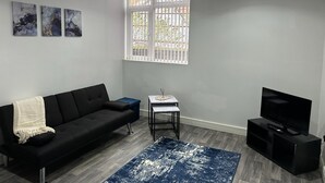 A living room with a black sofa, a small table, a TV, and a window with bars. - Broad Street, Wolverhampton - 2 Bedroom Apartment (Wolverhampton)