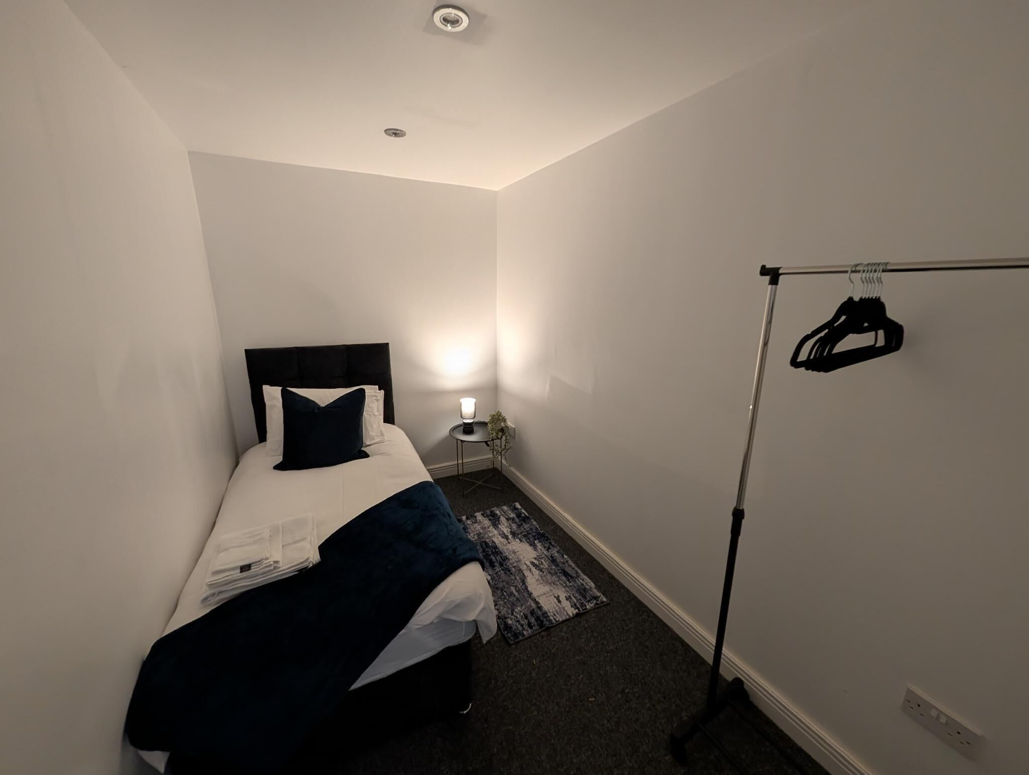 A small, neatly arranged bedroom with a bed, bedside lamp, and a clothing rack.