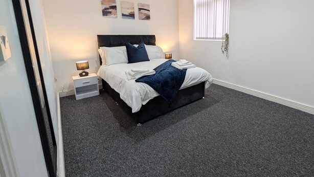 A modern bedroom with a bed, bedside tables, and a window with blinds. - Broad Street, Wolverhampton - 2 Bedroom Apartment (Wolverhampton)