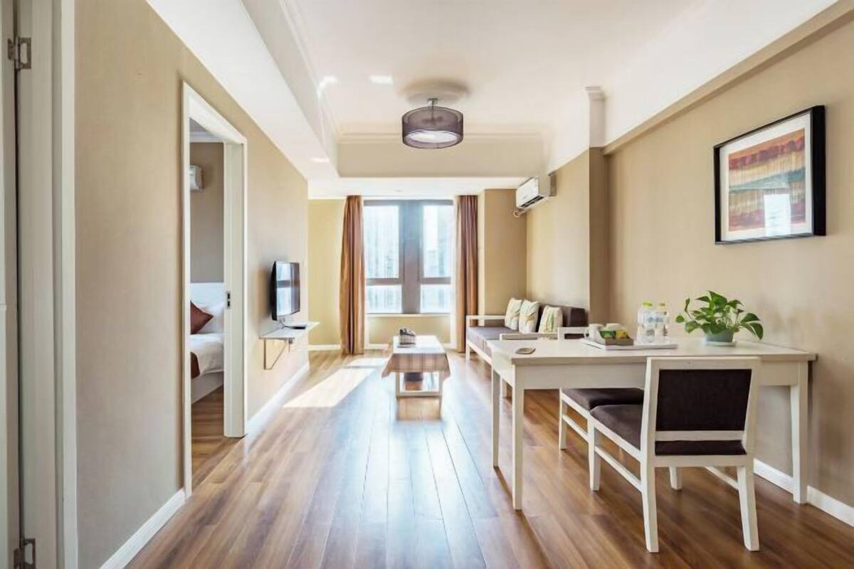 Una habitación de hotel con suelo de madera, una mesa de comedor con sillas, un sofá, un televisor y una ventana con cortinas.