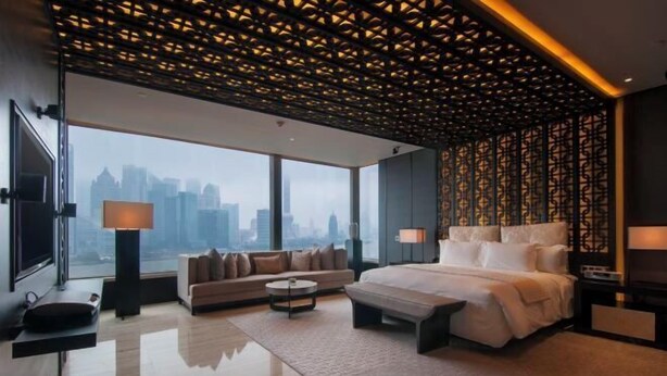 Ein modernes Hotelzimmer mit einem großen Bett, einer Couch, einem Couchtisch und Blick auf die Stadt durch große Fenster.