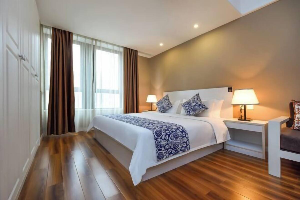 Un dormitorio con una cama grande, dos mesitas de noche con lámparas, suelo de madera y una ventana con cortinas.