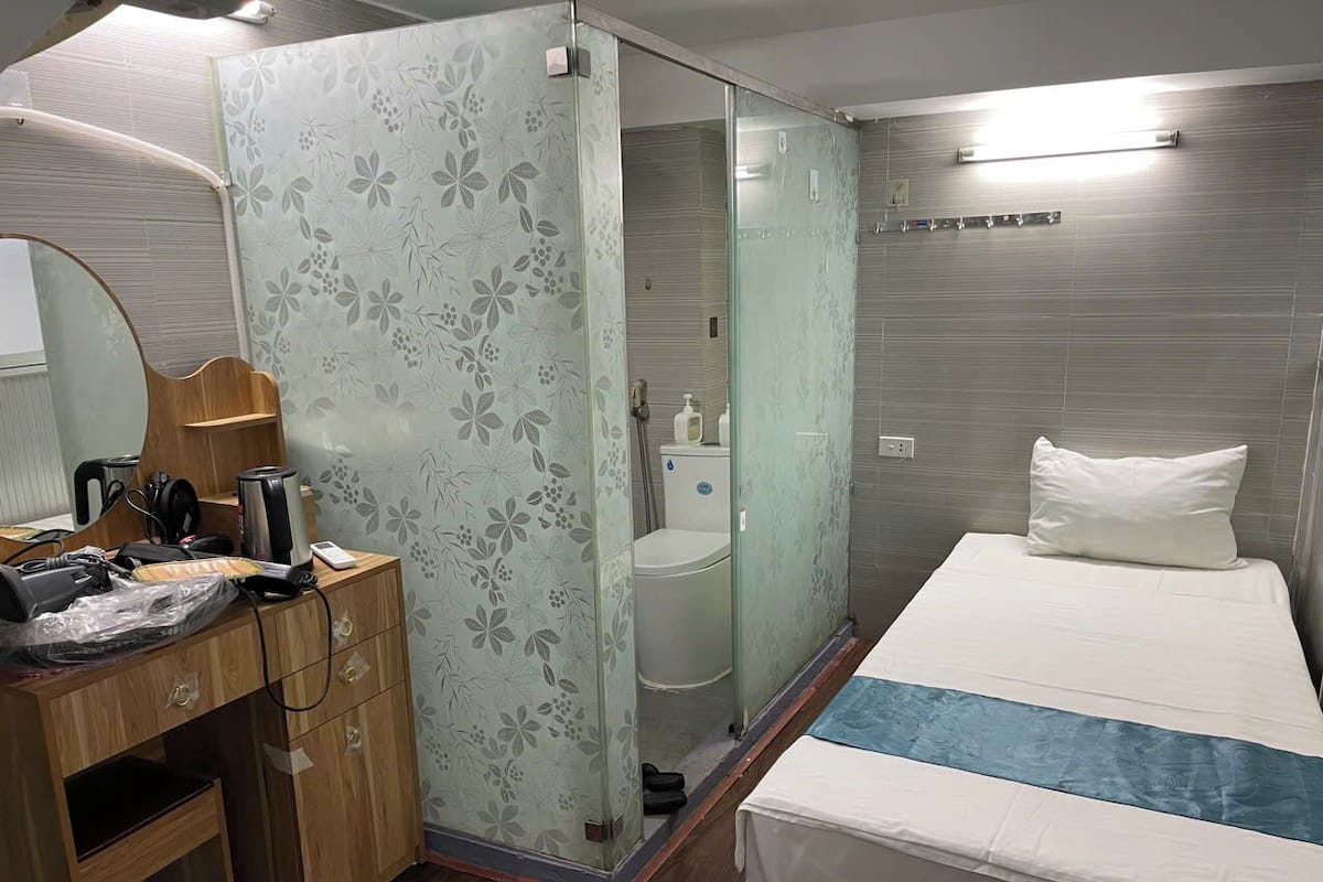 Habitación de hotel con un escritorio de madera, una cama con colcha azul y blanca, cabina de ducha de vidrio y pared con azulejos grises.