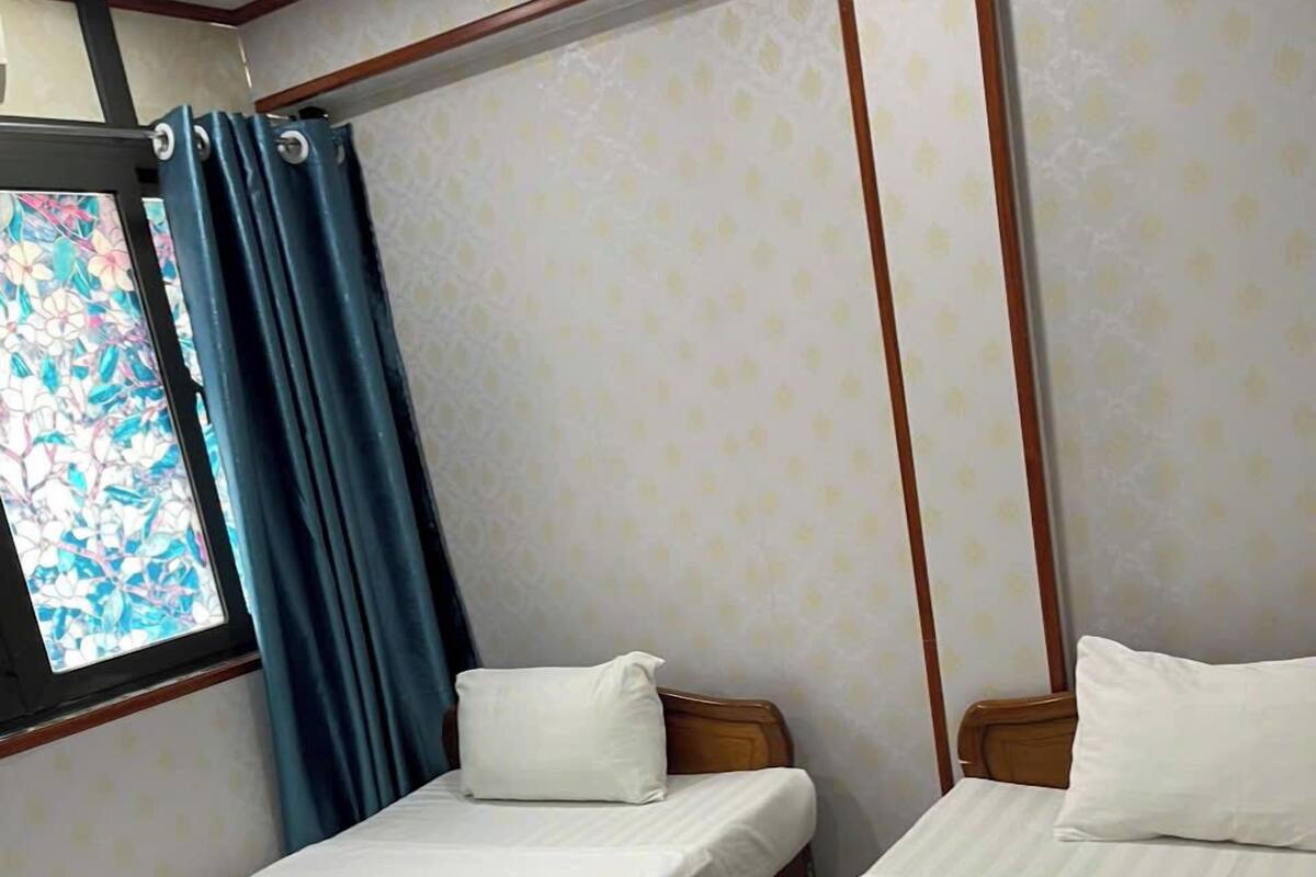 Una habitación con dos camas, cada una con colchón blanco y cubrecama azul. Hay una ventana con cortinas azules, y las paredes tienen un papel tapiz estampado.