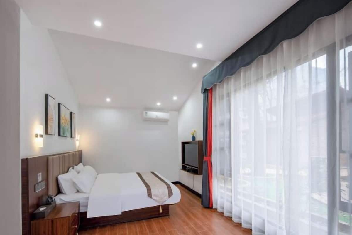 Habitación de hotel con cama, mesita de noche, televisor y ventanas amplias con cortinas translúcidas.