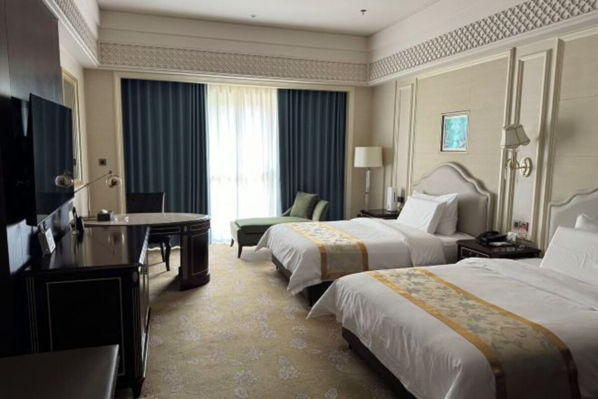 Habitación de hotel con dos camas, un escritorio, una silla, un televisor y un ventanal con cortinas.