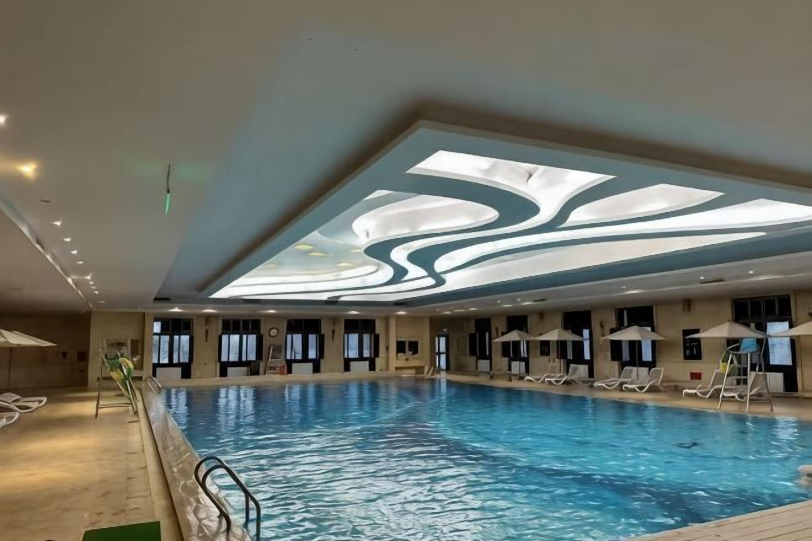 A large indoor swimming pool with a ceiling design, sun lounger area, and a poolside bar.