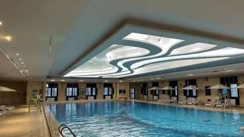 A large indoor swimming pool with a ceiling design, sun lounger area, and a poolside bar.