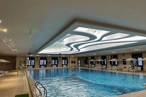 Una amplia piscina cubierta con diseño en el techo, zona de sillones de sol y un bar junto a la piscina.