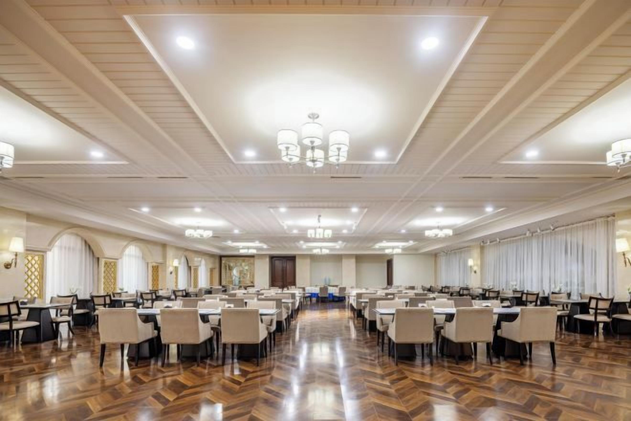 A large banquet hall with rows of tables and chairs, high ceilings with chandeliers, and a polished wooden floor.