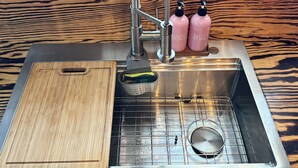 Ein Edelstahl-Spülbecken mit Schneidebrett aus Holz, einem Metallsieb und zwei rosa Seifenspendern mit den Aufschriften 'HANDS' und 'DISHES'.