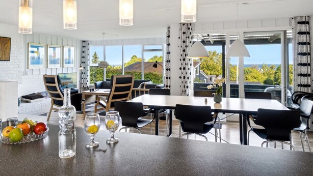 Un espace de salle à manger moderne, doté d’une grande table, de chaises et de luminaires suspendus. Des tableaux encadrés ornent le mur, et de grandes fenêtres offrent une vue sur l’extérieur.