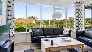 Un salon moderne avec un canapé en cuir noir, une table basse en bois et un téléviseur. De grandes fenêtres offrent une vue sur l’extérieur.