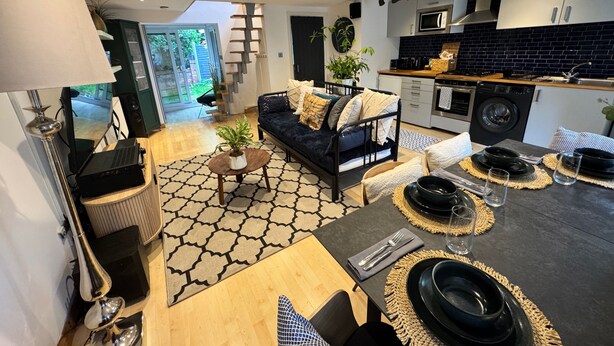 A modern kitchen with a microwave, oven, and sink. There is a dining area with a table set for two, a sofa, and a coffee table. A staircase leads to a balcony with a view of greenery. - Coventry Cozy Cottage (Coventry)