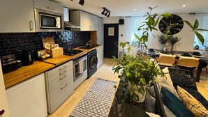 A modern kitchen with a black tiled backsplash, stainless steel appliances, and a wooden countertop. There is a dining area with a black clock and potted plants. - Coventry Cozy Cottage (Coventry)