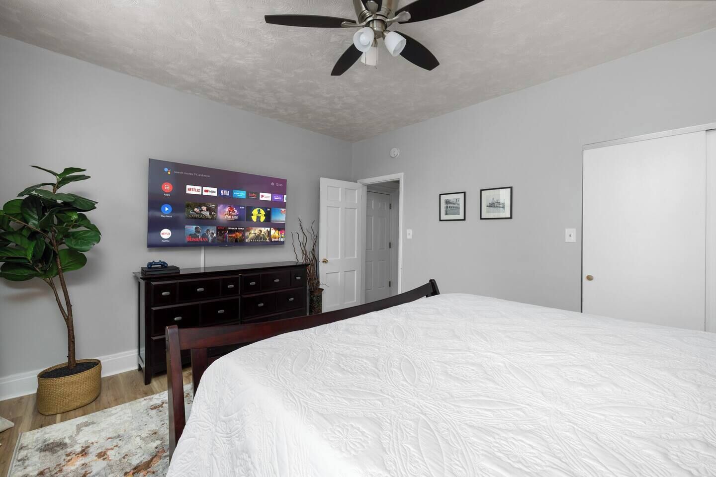 Een slaapkamer met een bed, een plafondventilator, een aan de muur bevestigde televisie, een dressoir en een deur.