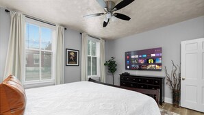 A bedroom with a bed, a ceiling fan, a TV on the wall, and a window with curtains.