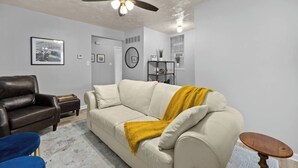 A living room with a beige sofa, a dark brown leather chair, a blue chair, a wooden side table, a small round wooden table, a ceiling fan, and a framed picture on the wall.