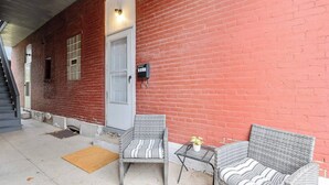 A covered outdoor seating area with wicker furniture and a small table, adjacent to a red brick building with a white door and a mailbox.