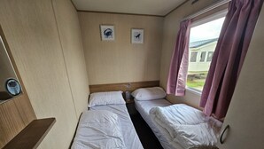 Une petite chambre à coucher, de taille modeste, avec deux lits simples, une tête de lit en bois, une fenêtre avec des rideaux et deux tableaux encadrés accrochés au mur.