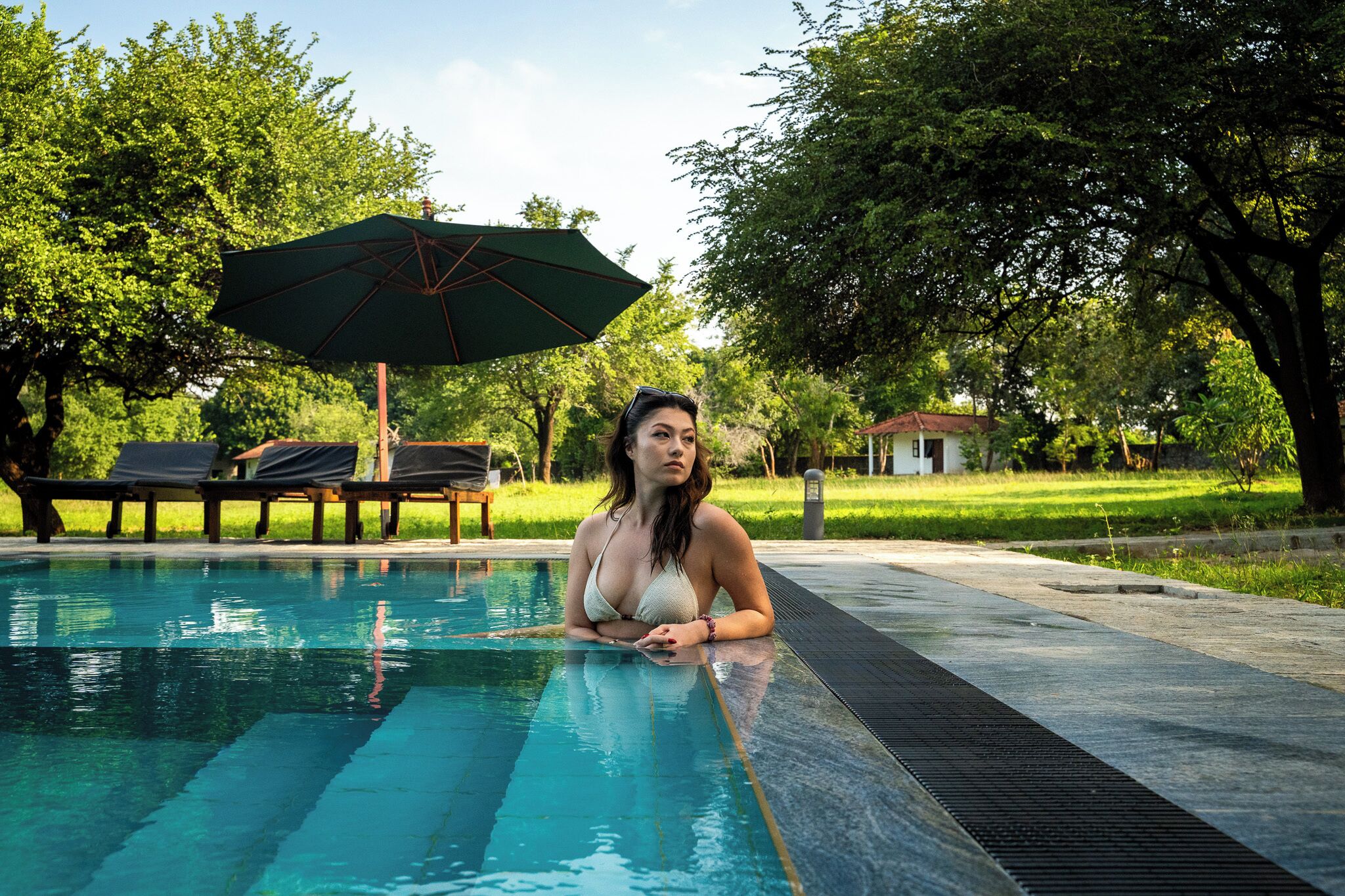 A woman in a swimming pool with a green umbrella and lounge chairs in the background.